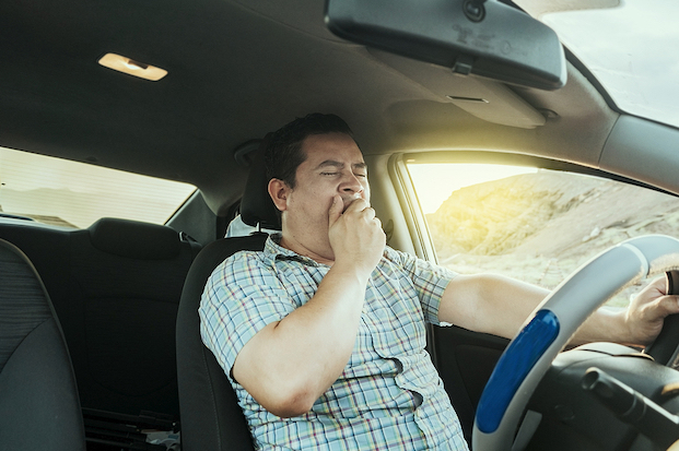 Caucasian man sitting behind steering wheel driving, while holding the steering wheel with his left hand, yawning and holding up his right hand over his mouth, with his eyes closed.